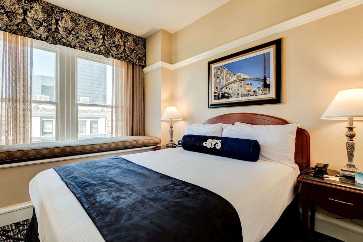 Ein Hotelzimmer mit einem großen Bett, einem Fenster mit Vorhängen, einem gerahmten Bild an der Wand und einem Nachttisch mit einer Lampe.