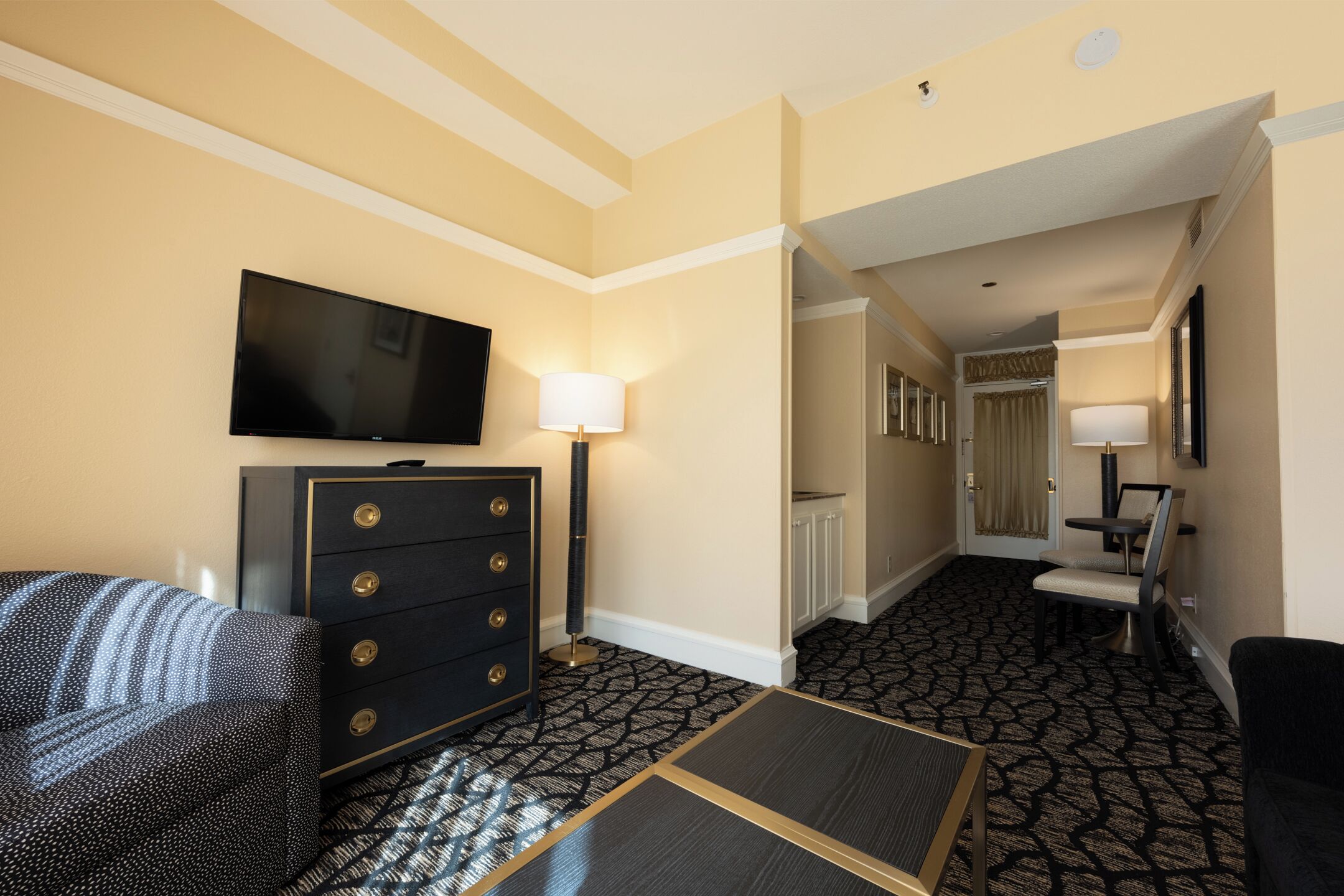 A hotel room with a flat-screen TV, a dresser, a lamp, a patterned carpet, and a seating area with a chair and a small table.