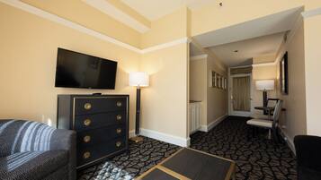 A hotel room with a flat-screen TV, a dresser, a lamp, a patterned carpet, and a seating area with a chair and a small table.