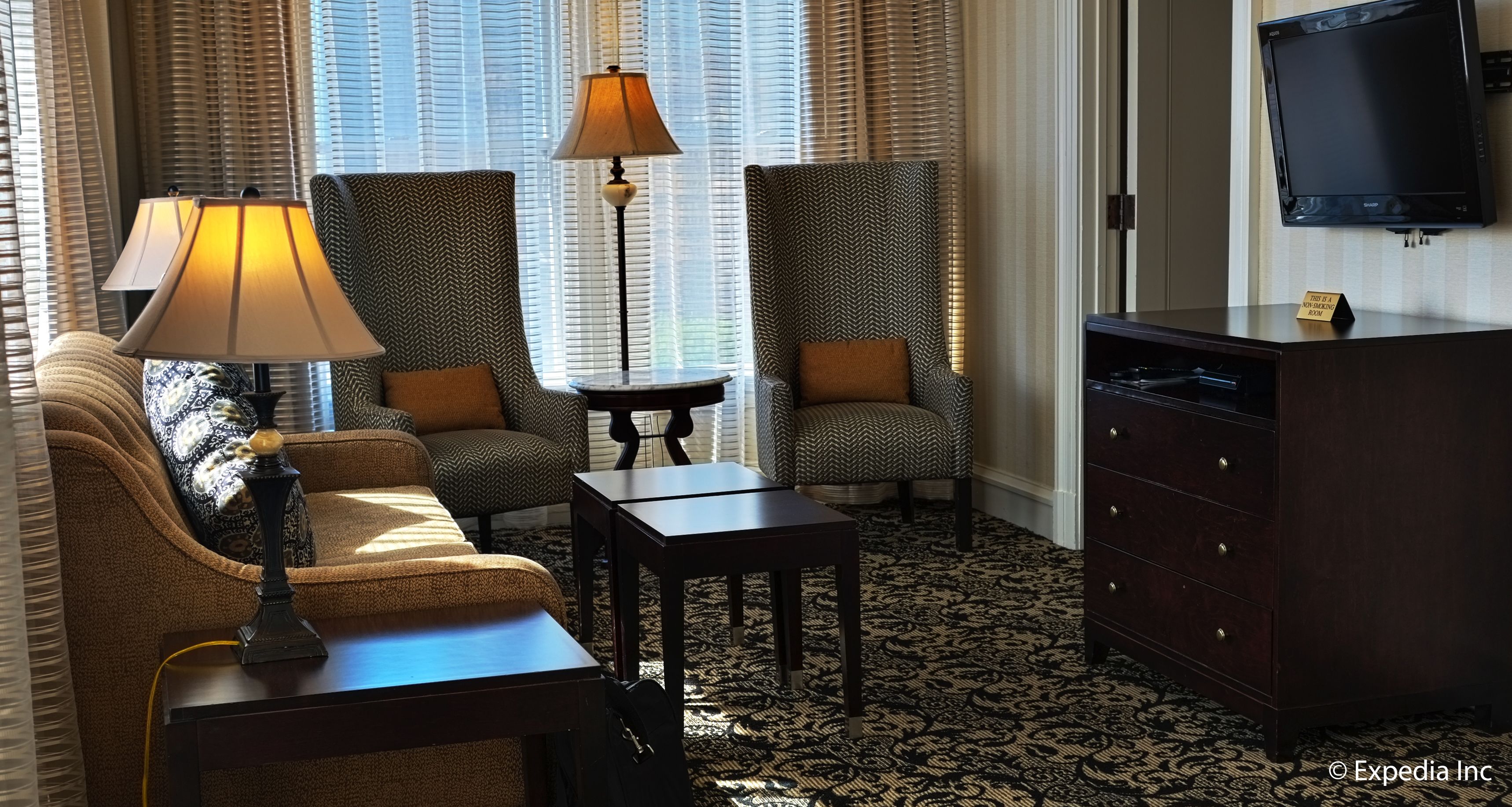 Ein Hotelzimmer mit einer Couch, zwei Sesseln, einem kleinen Tisch, einer Lampe, einem Fernseher und einem Schminktisch.