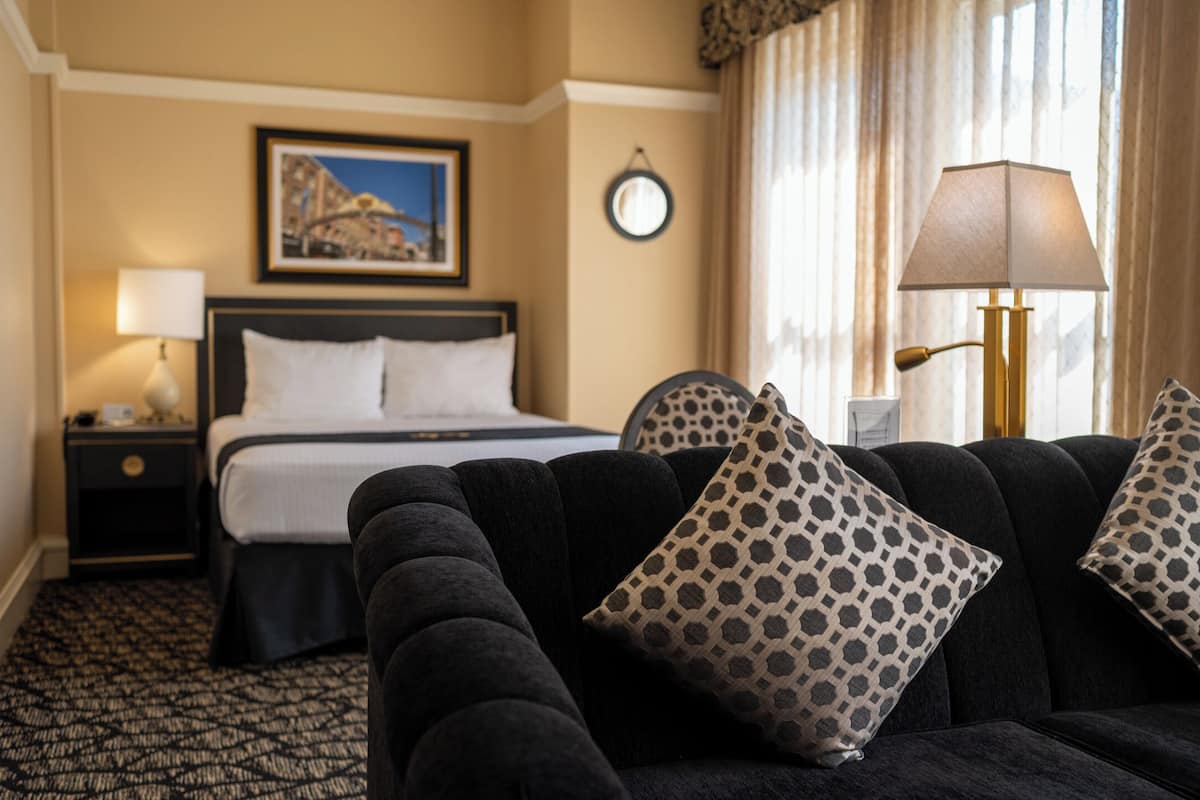 Ein Hotelzimmer mit Bett, Nachttisch, Lampe und einer Couch mit gemusterten Kissen.