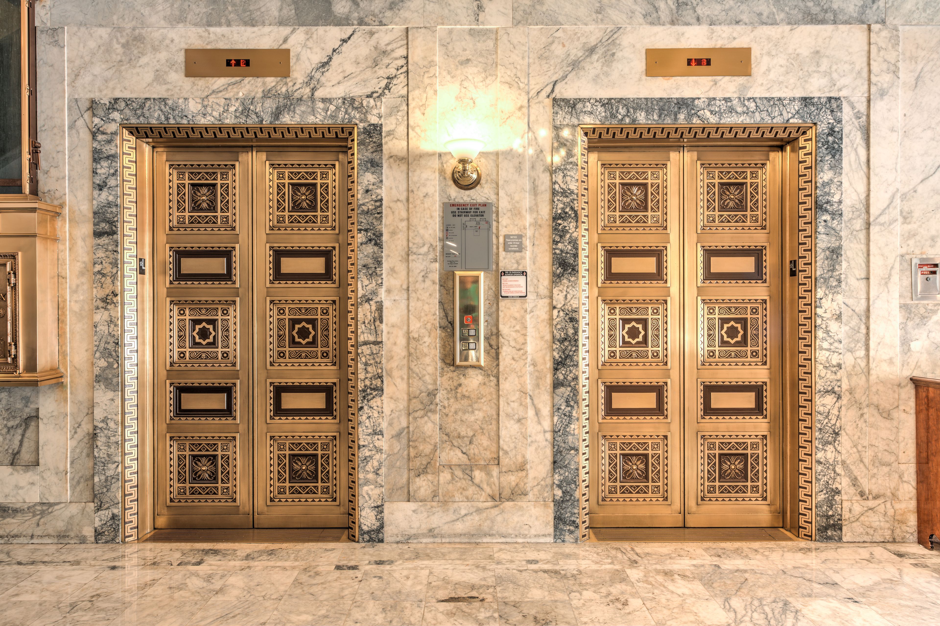 Two elevator doors with intricate designs, a marble wall, and a light fixture above the doors.