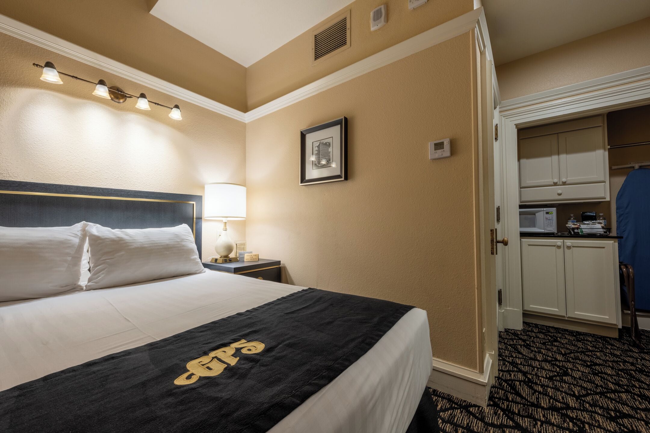 Habitación de hotel con una cama grande, una lámpara de noche, un cuadro en la pared y una pequeña cocina.