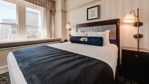 Una habitación de hotel con una cama grande, una ventana con persianas, una mesita de noche con lámpara y un cuadro en la pared.