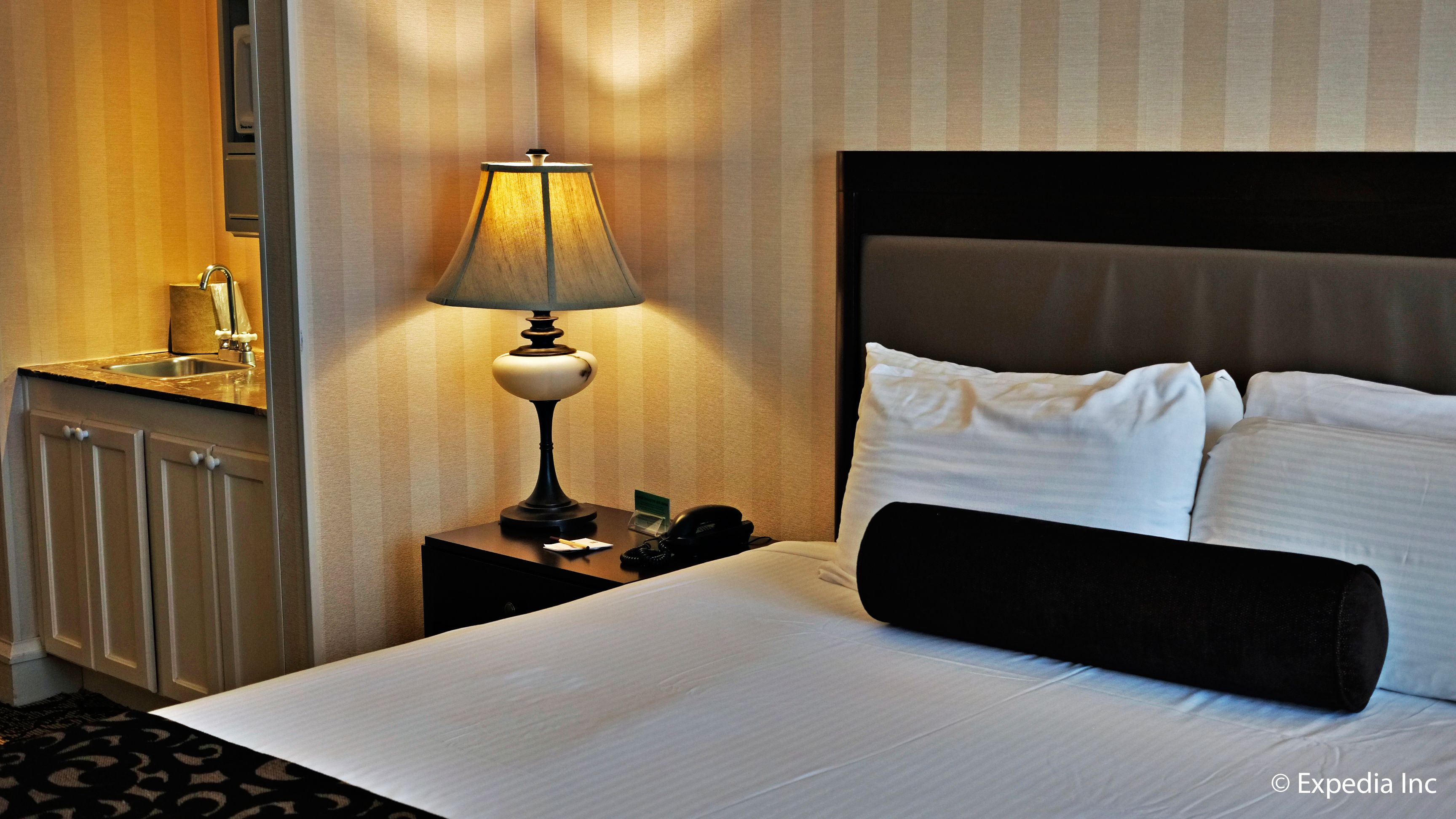 A hotel room with a bed, a nightstand with a lamp, a telephone, and a small table with a card and a pen.