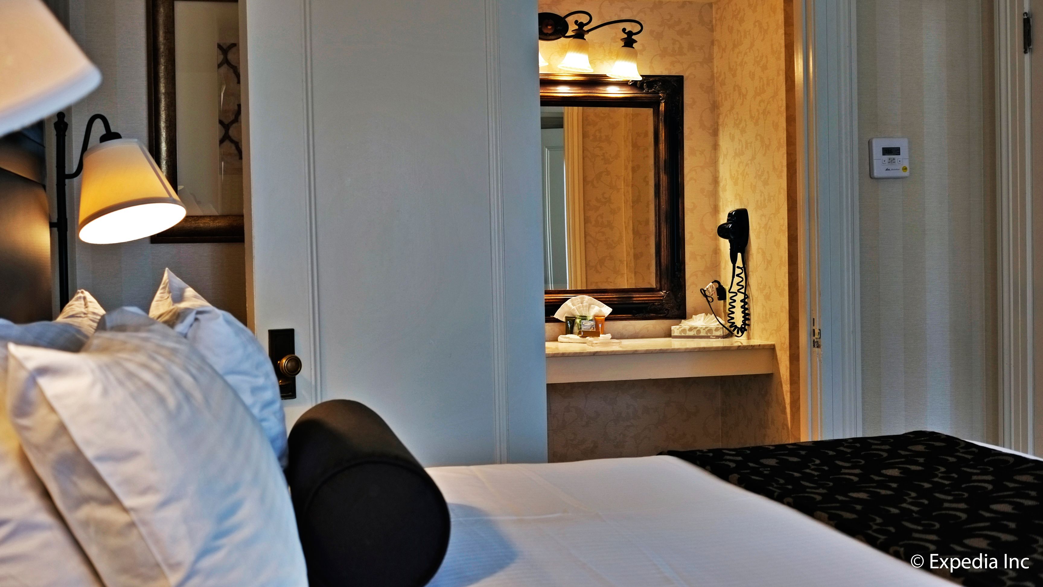 Une chambre d’hôtel avec un lit, une lampe de chevet, une table de nuit, un miroir et une salle de bain visible à travers une porte ouverte.