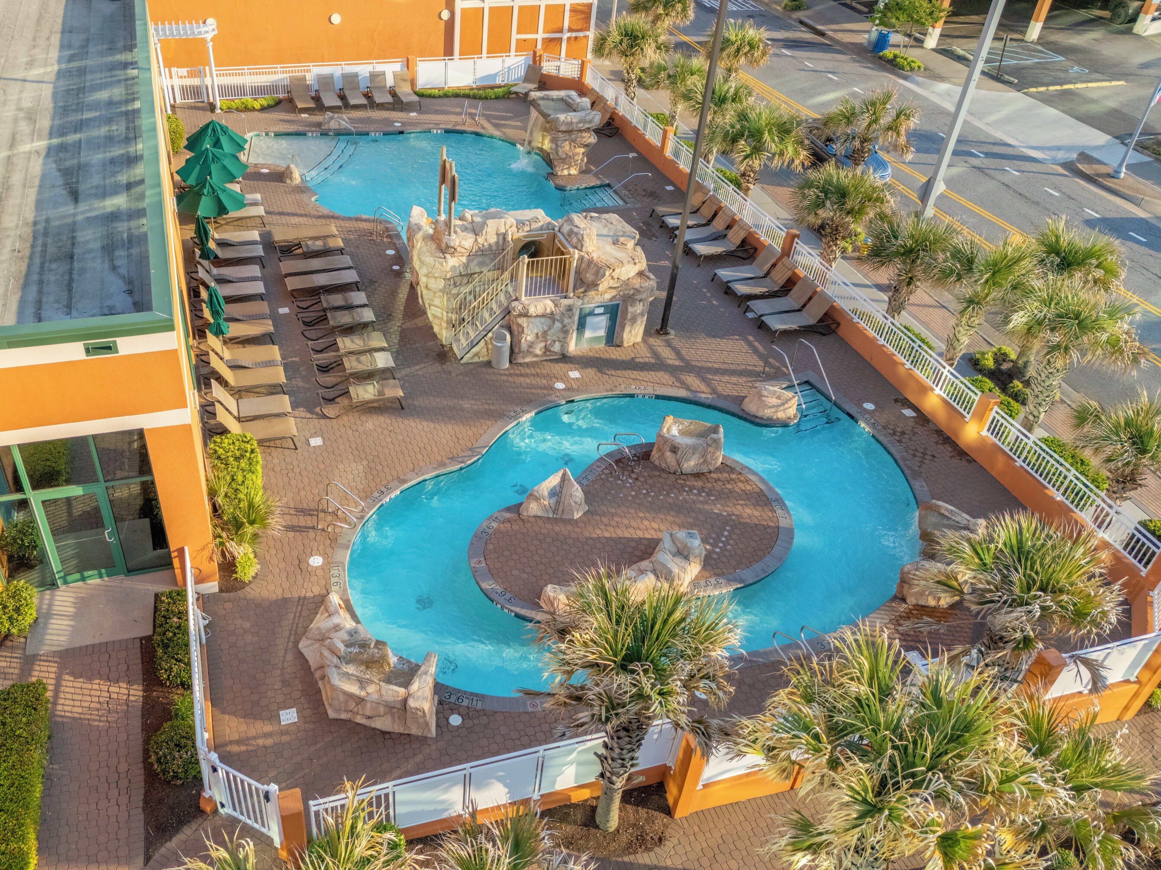 Un área de piscina en la azotea con varias piscinas, sillas de descanso y palmeras.