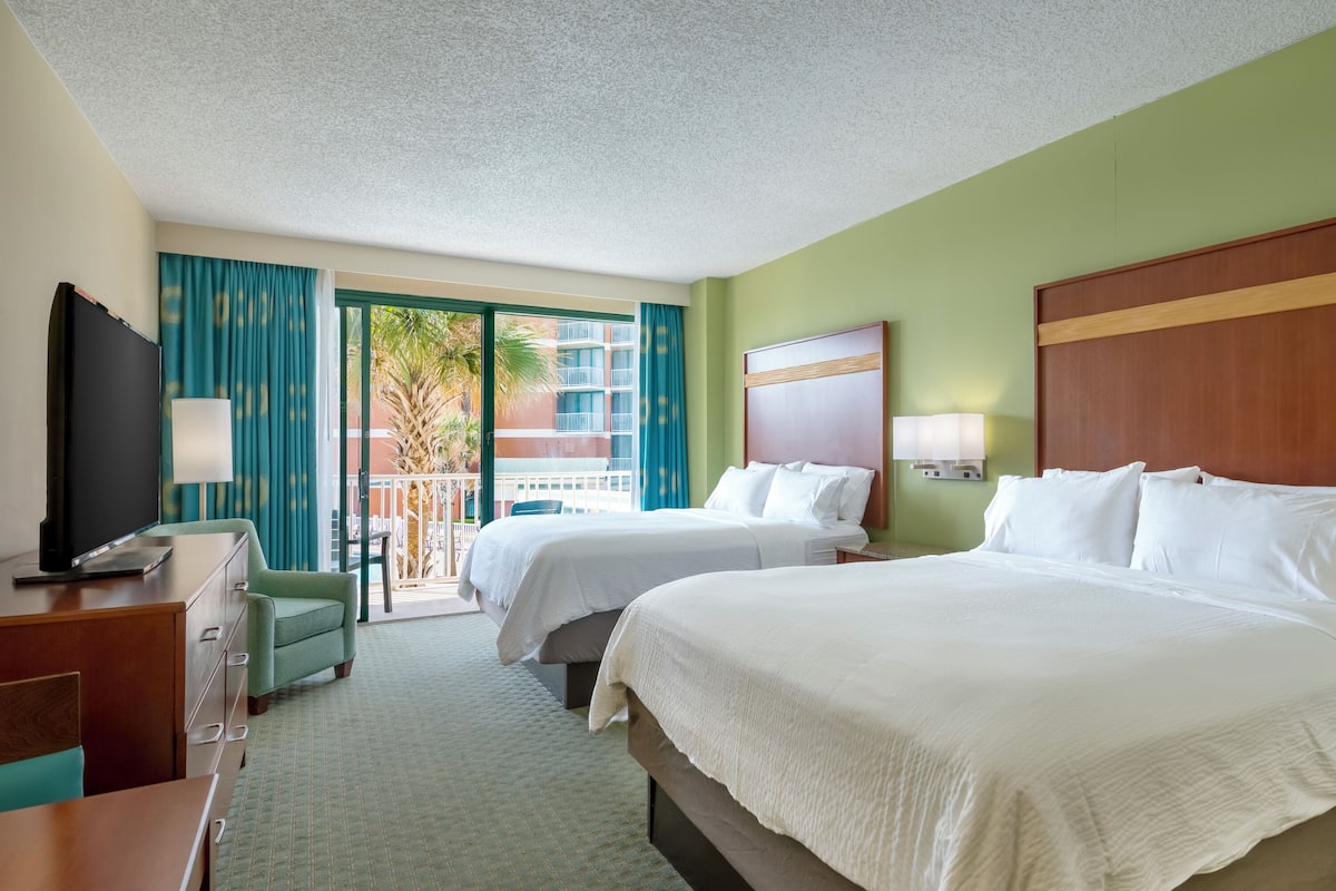 A hotel room with two beds, a television, a desk, and a view of a balcony with palm trees.