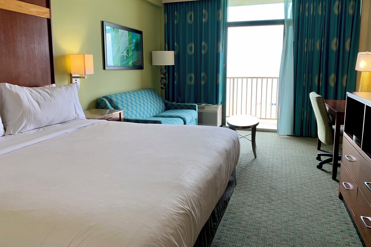 A hotel room with a large bed, a desk, a chair, a small table, a lamp, a picture on the wall, and a view through the window.