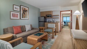 Una sala moderna con sofá, mesa de centro y sillas, una cocina con microondas y refrigerador, y vistas a través de una puerta abierta.