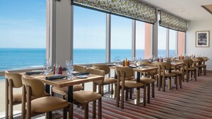 Un comedor con mesas y sillas, ventanas amplias con vistas al océano, y una alfombra a rayas.