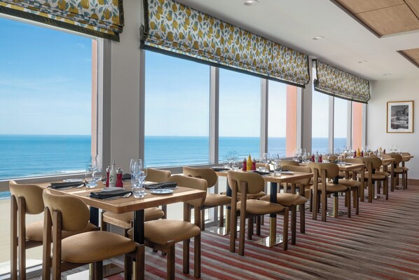 A dining area with tables and chairs, large windows with ocean views, and a striped carpet. - Holiday Inn & Suites Virginia Beach North Beach by IHG (Virginia Beach)