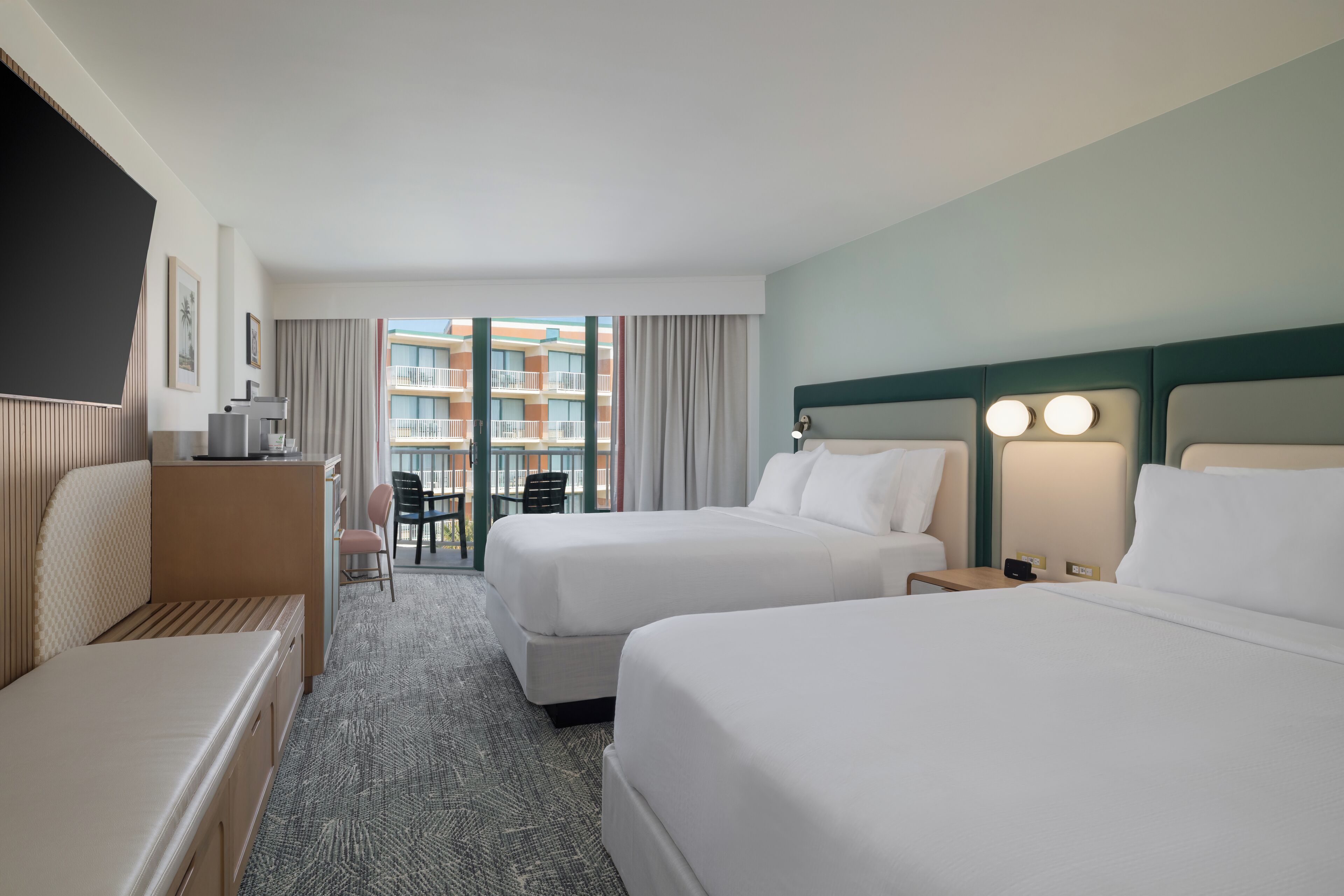 A hotel room with two beds, a desk, a chair, and a view of a building with balconies.