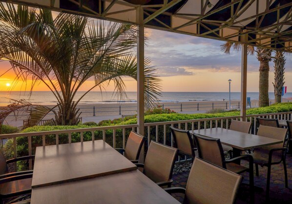 An outdoor seating area with tables and chairs, overlooking a beach and ocean at sunset. - Holiday Inn & Suites Virginia Beach North Beach by IHG (Virginia Beach)