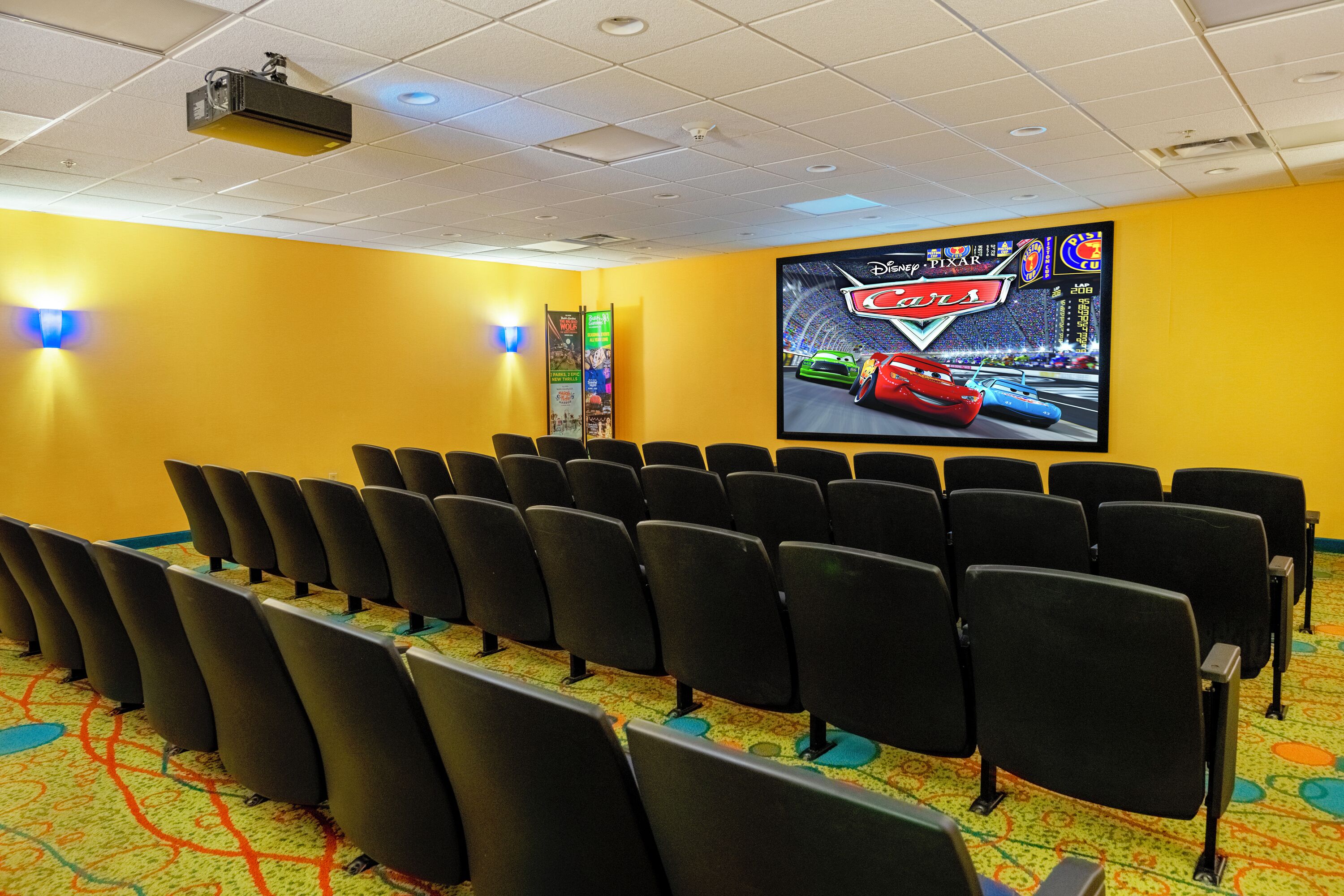 A movie theater with rows of black seats, a large screen displaying a Disney Pixar Cars movie, and a colorful carpet.