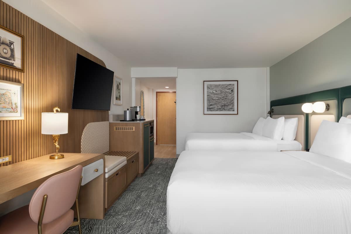 A hotel room with a bed, desk, chair, kitchenette, and a TV mounted on the wall.