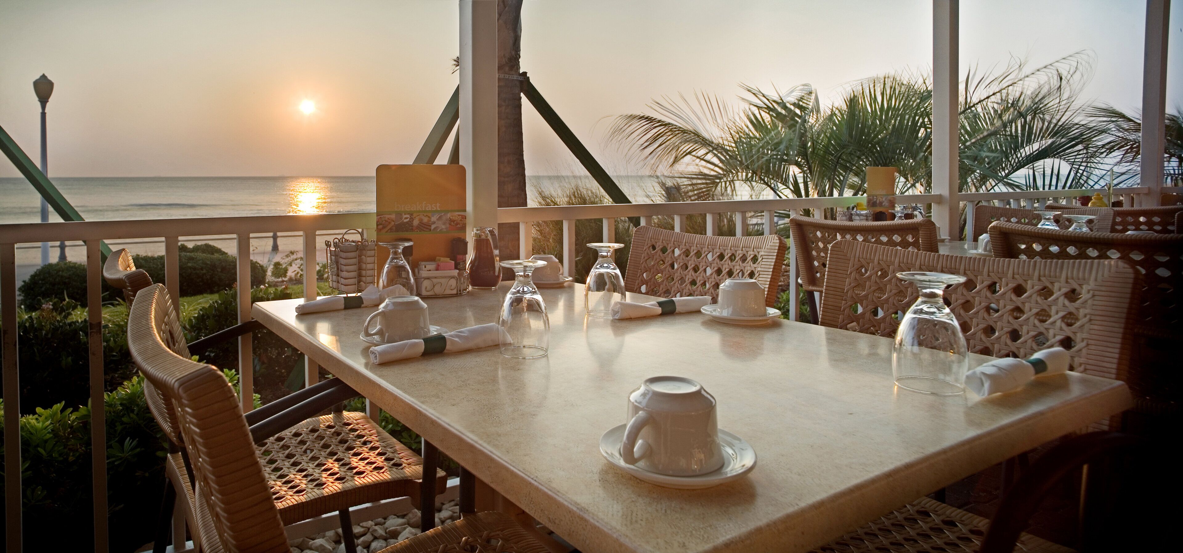 A dining table set for breakfast with a view of the sea and palm trees.