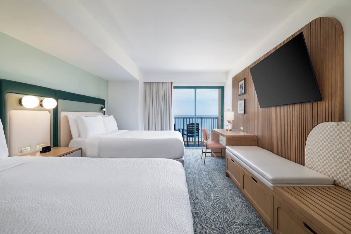 A hotel room with two beds, a desk, a chair, a TV, and a balcony with a view.