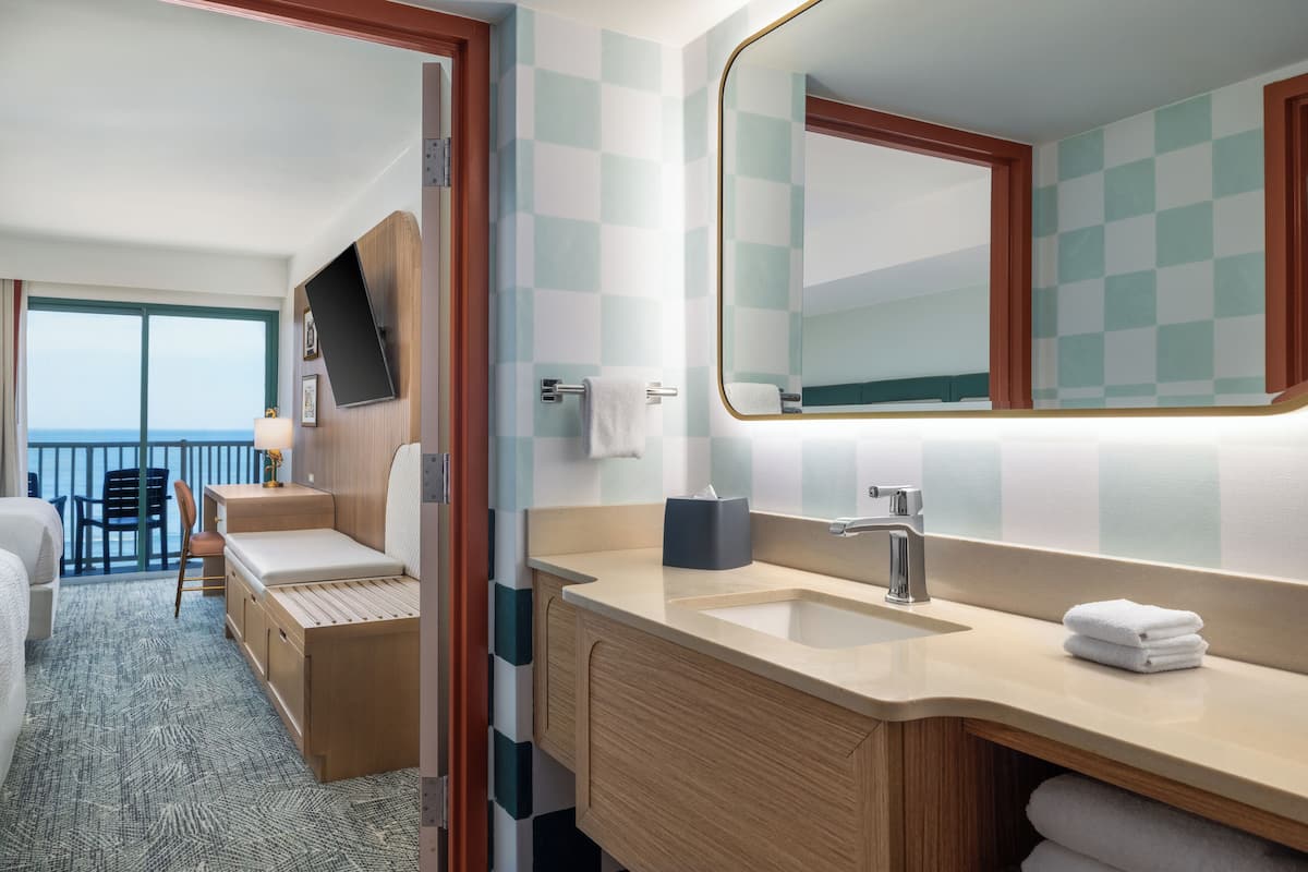 A hotel room with a balcony, a bed, a sitting area, a bathroom with a sink and mirror, and a view of the sea.