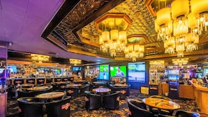 Interior de un casino con varias mesas de juego, zona de bar y pantallas grandes mostrando partidas.