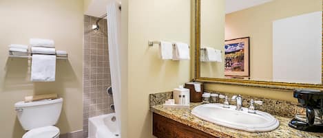 A bathroom with a toilet, sink, and shower. There are towels on the wall and a framed picture above the mirror.