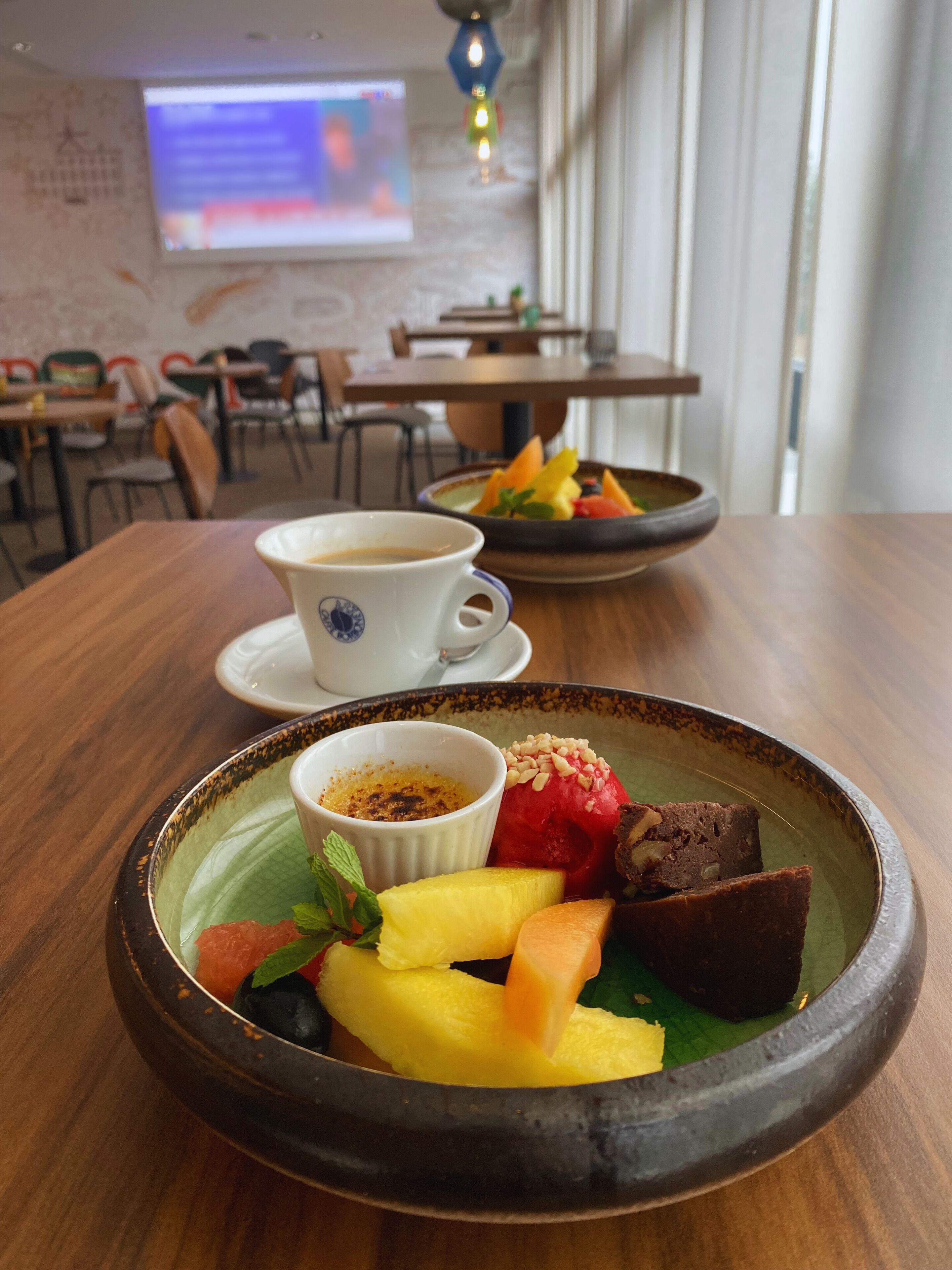 Una mesa de comedor con un plato de postre que contiene fruta y un recipiente pequeño de salsa, una taza de café y una pantalla de presentación al fondo.