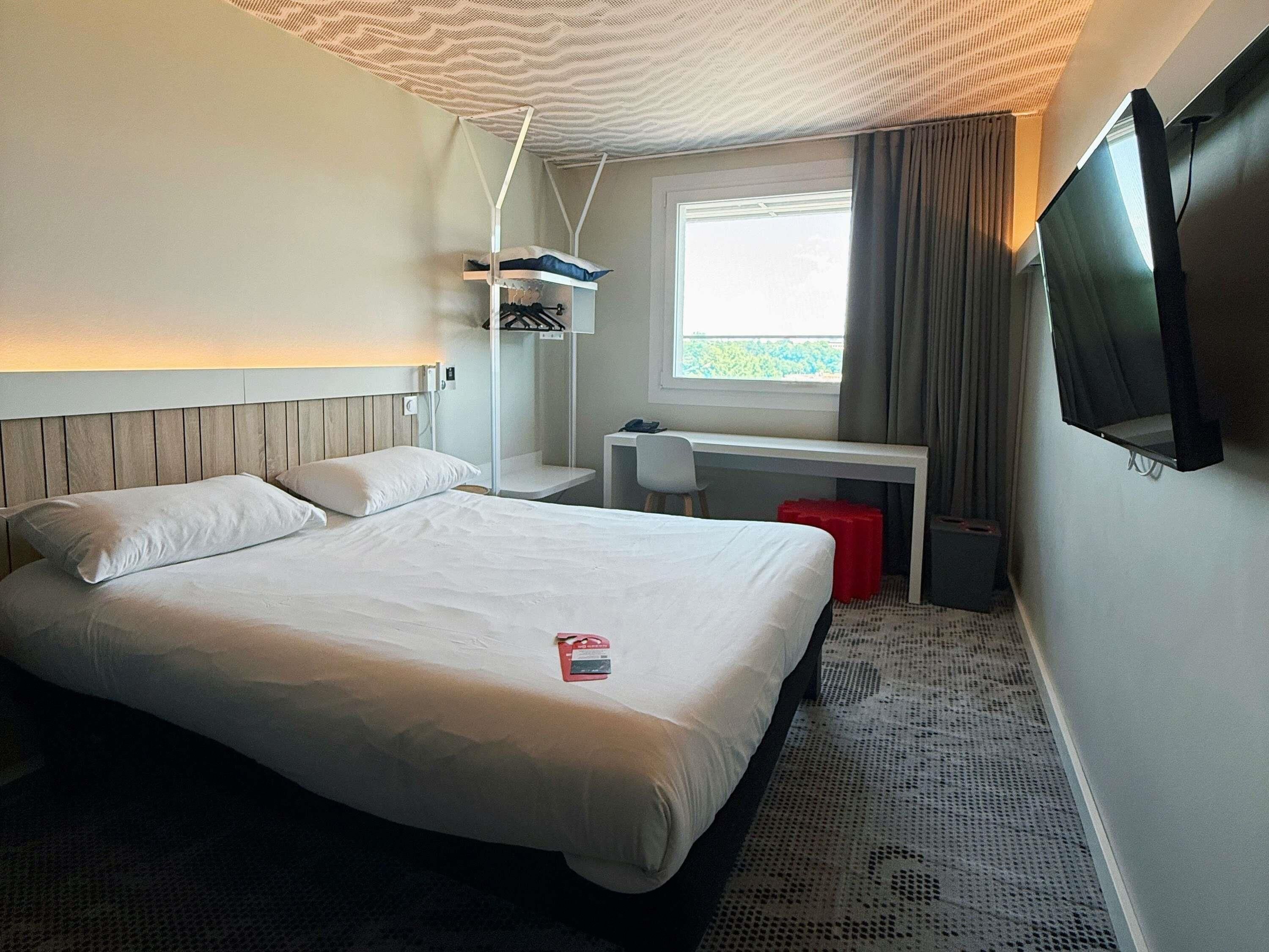 Habitación de hotel con cama, escritorio, televisor y ventana con cortinas.