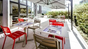 An outdoor seating area with tables and chairs, umbrellas, and a building with large windows in the background.