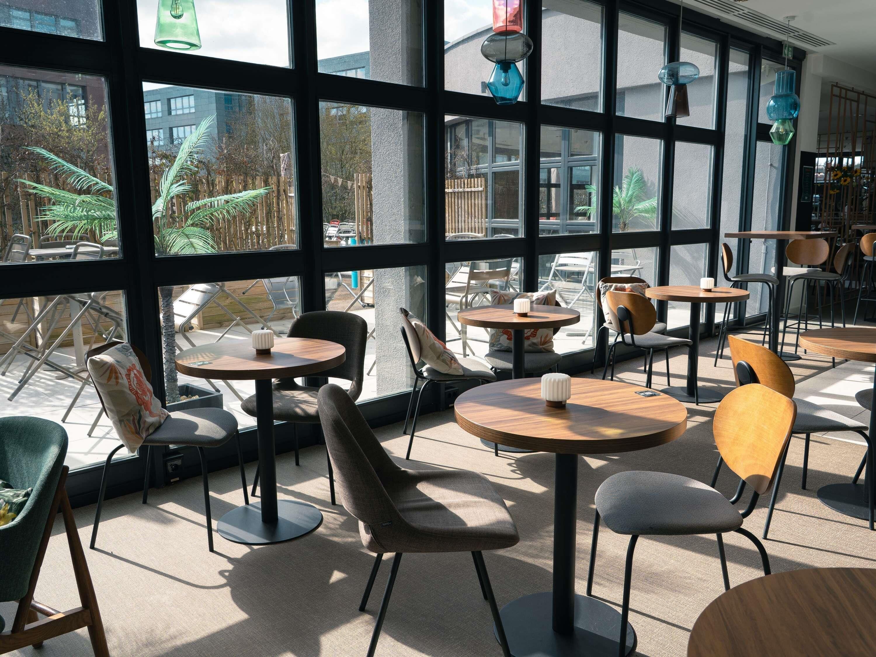 A modern indoor seating area with round tables and chairs, large windows, and hanging pendant lights.
