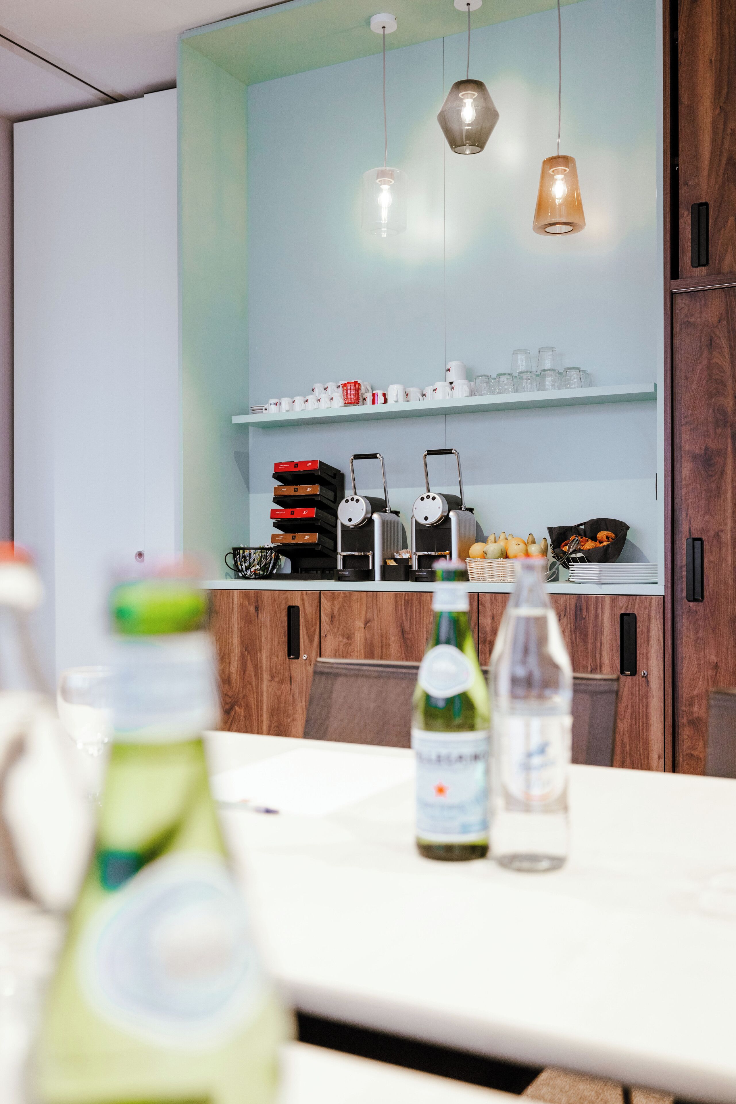 Une cuisine moderne avec des meubles en bois, un plan de travail blanc et des luminaires suspendus. On peut voir des bouteilles sur le plan de travail et une machine à café sur l’étagère.