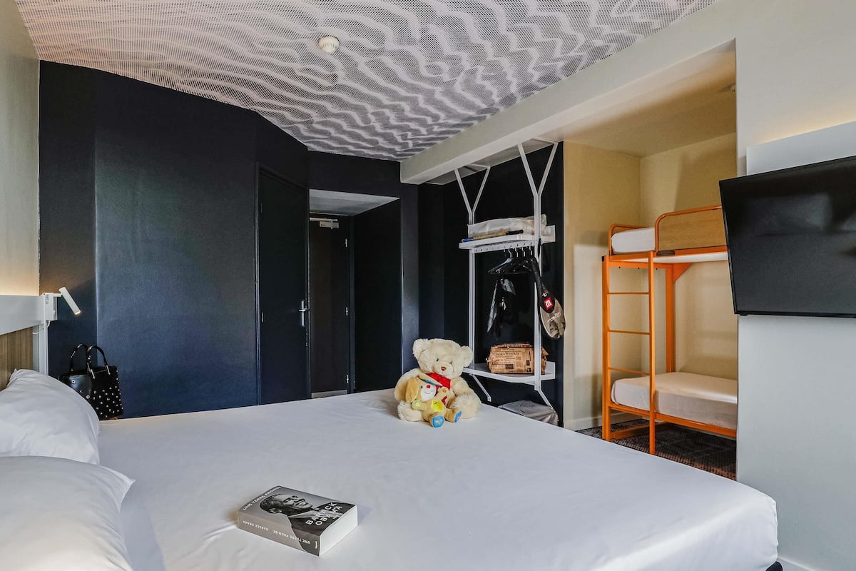 A hotel room with a bed, a teddy bear, a book, a coat rack, and a TV on the wall.