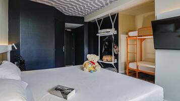 Une chambre d’hôtel avec un lit, un ours en peluche, un livre, un porte-manteau et un téléviseur fixé au mur.