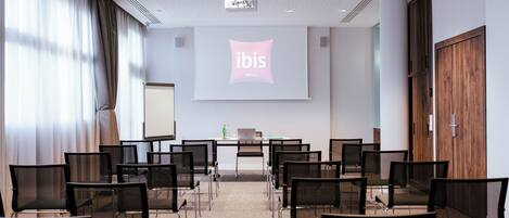 Un salón de conferencias con una pantalla de proyector mostrando el logotipo de ibis Hotels, sillas dispuestas frente a una mesa con una computadora portátil, y una puerta de madera a la derecha.