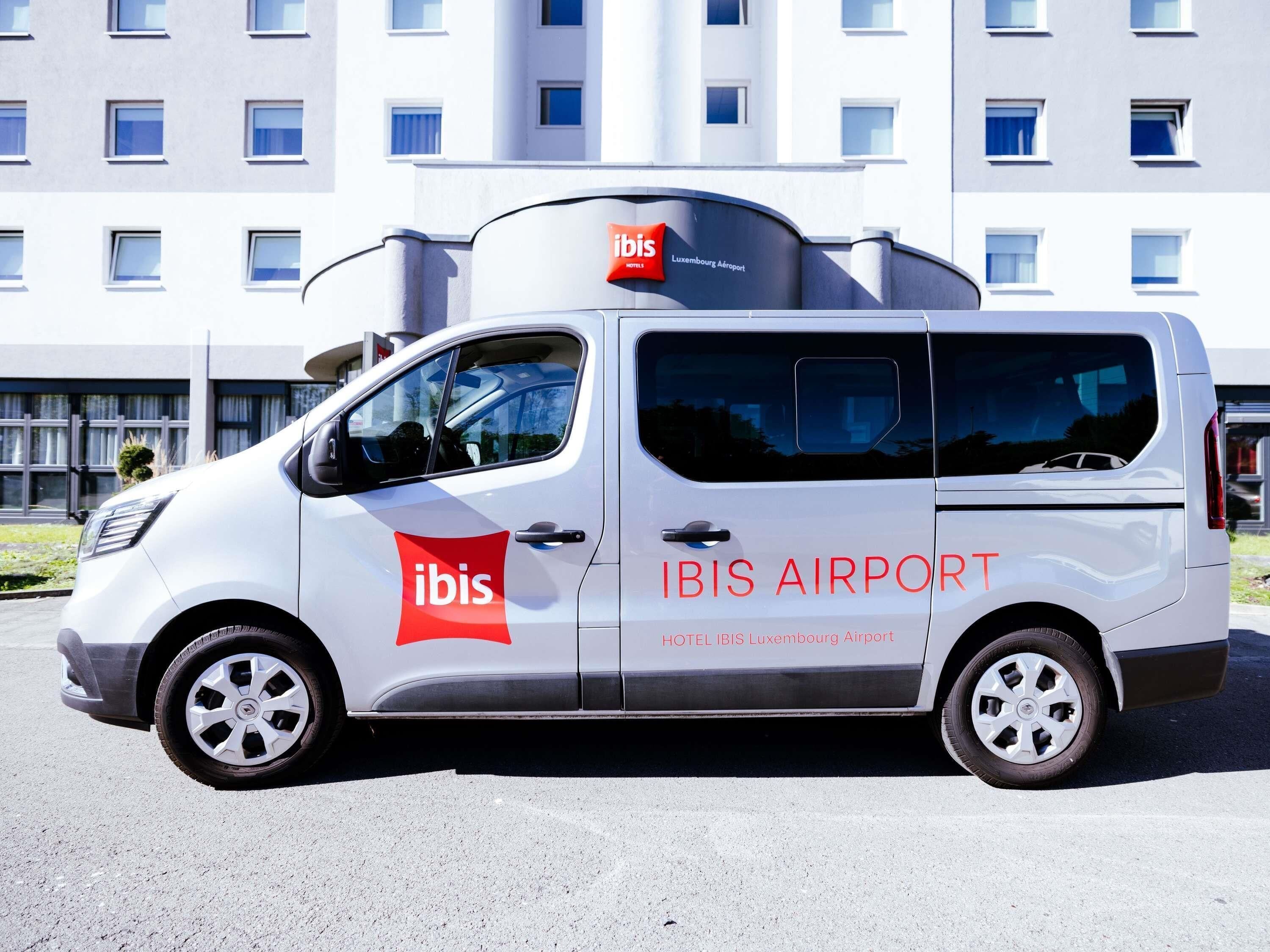 Una furgoneta blanca amb el logotip 'ibis' i la inscripció 'HOTEL IBIS Luxembourg Airport', aparcada davant d'un edifici amb diverses finestres.