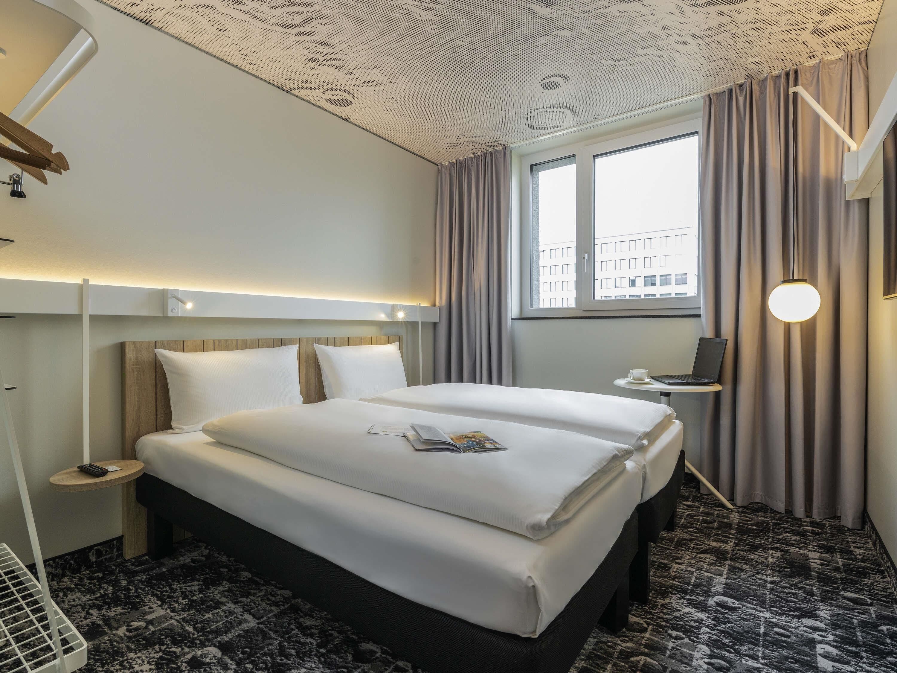 Een hotelkamer met een groot bed, nachtkastjes, een bureau met een laptop en een raam met gordijnen.