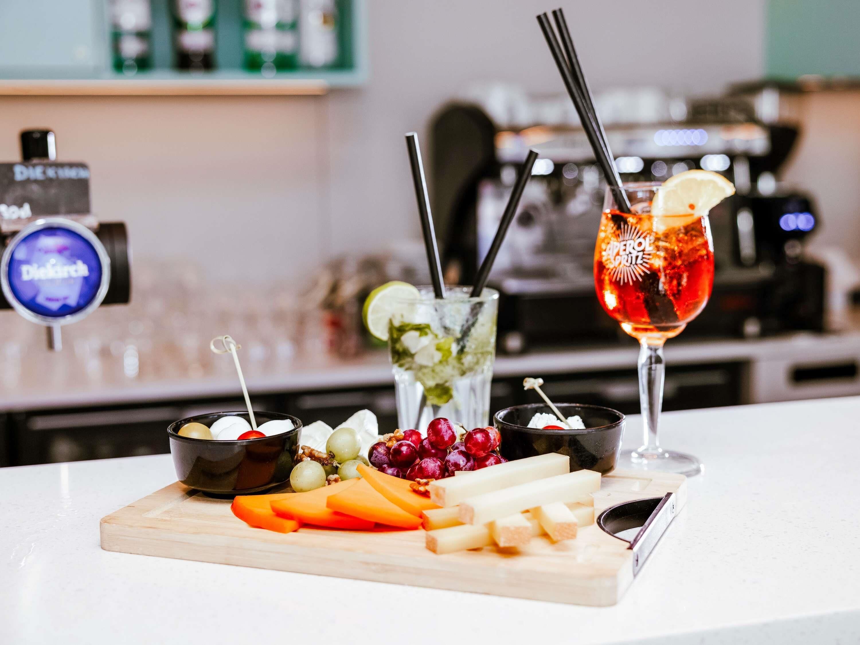 A bar area with a selection of drinks, including a cocktail with a lemon garnish, and a variety of fruits and cheeses on a wooden board.