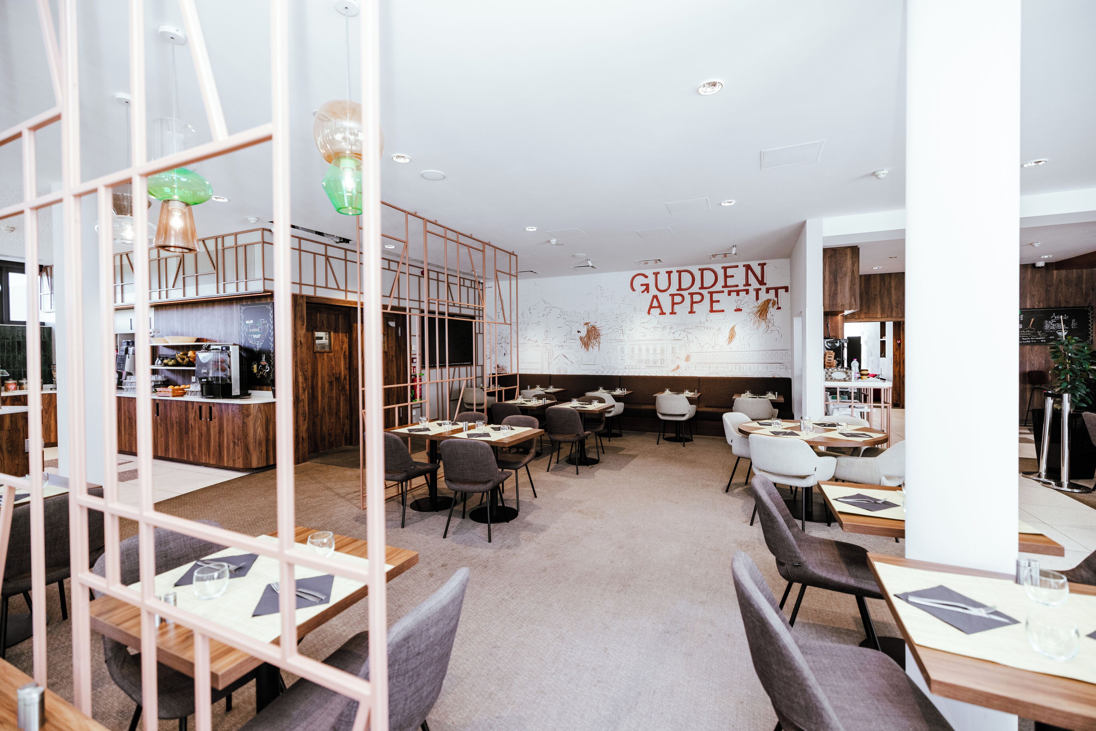 A modern restaurant with a high ceiling, wooden accents, and a central wall with the text "GUDDEN APPE". Tables are arranged for dining, and there are chairs and a counter area visible.