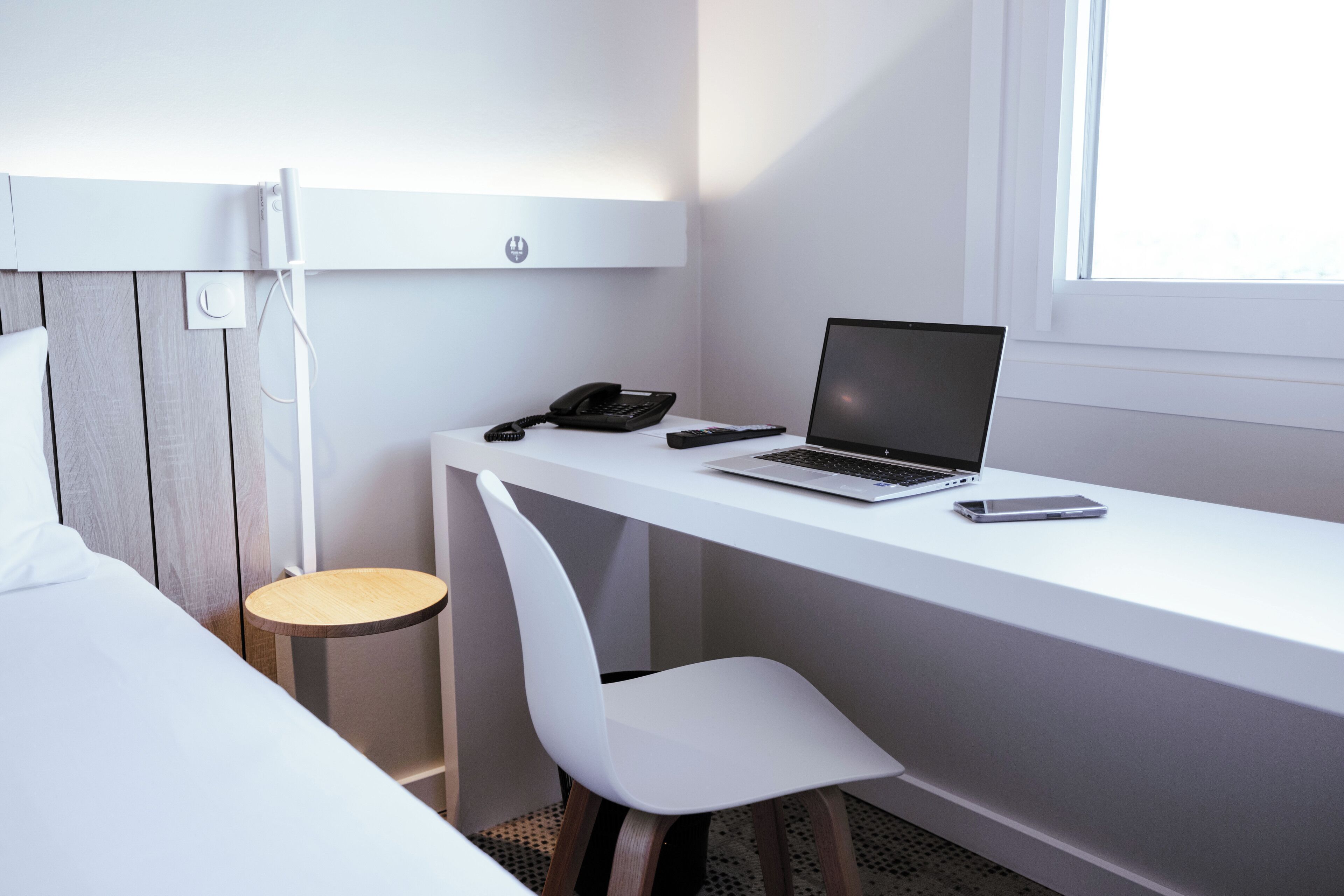 A modern hotel room with a white desk, a laptop, a phone, and a chair.