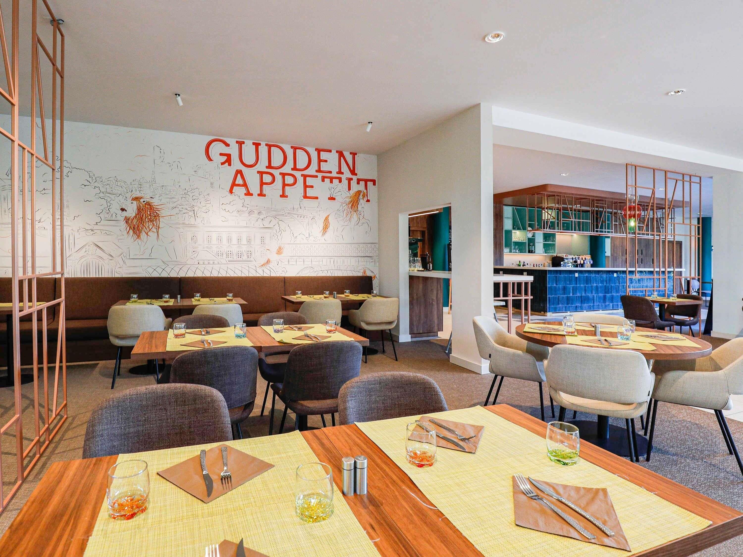 Un interior de restaurante con mesas dispuestas para comer, una zona de bar y una pared con un cartel de gran tamaño.