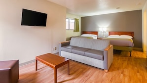 A hotel room with a sofa, a wooden table, a bed with a headboard, a flat-screen TV mounted on the wall, and a window with curtains. - Studio 6 Extended Stay - Greensboro, NC (Greensboro)