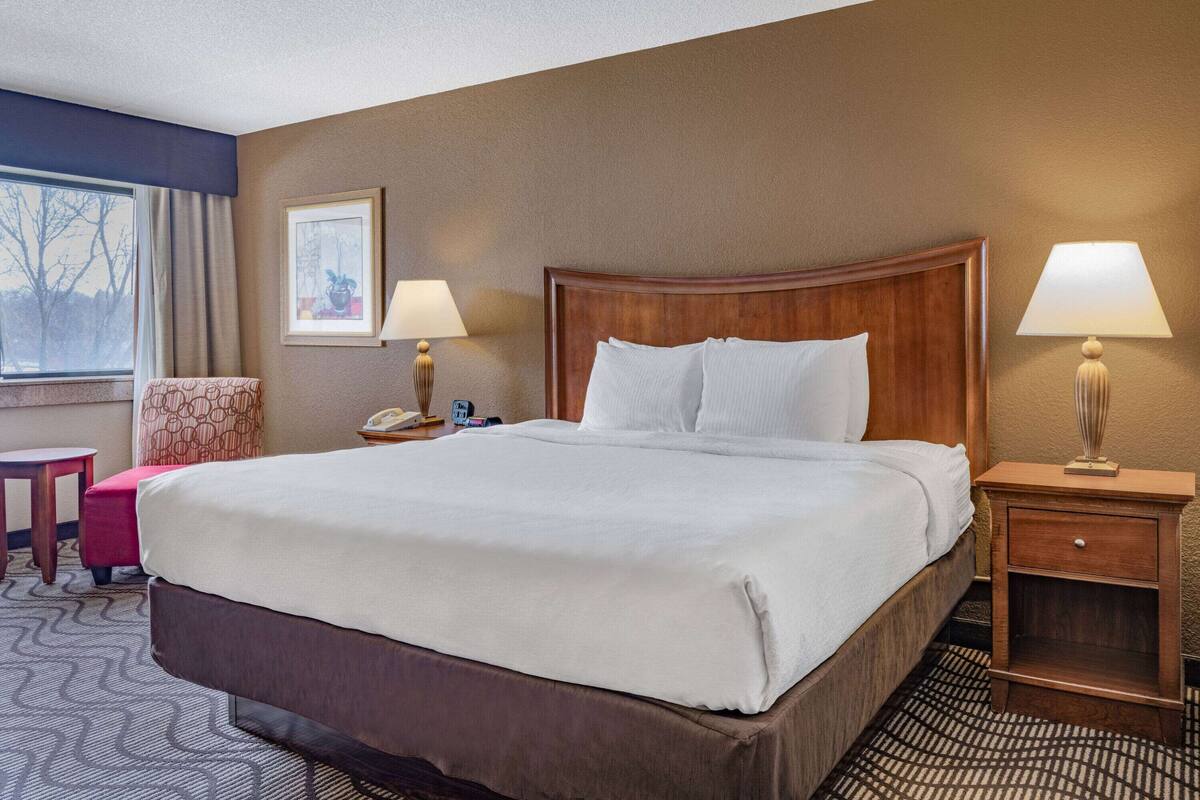 Une chambre d’hôtel avec un grand lit, deux tables de chevet avec des lampes, un petit bureau, une chaise et une fenêtre donnant sur des arbres.