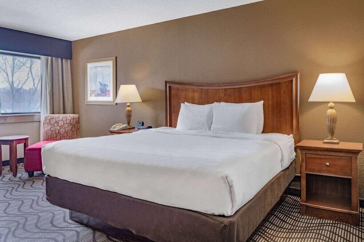 Une chambre d’hôtel avec un grand lit, deux tables de chevet avec des lampes, une chaise, un bureau et une fenêtre avec des rideaux.