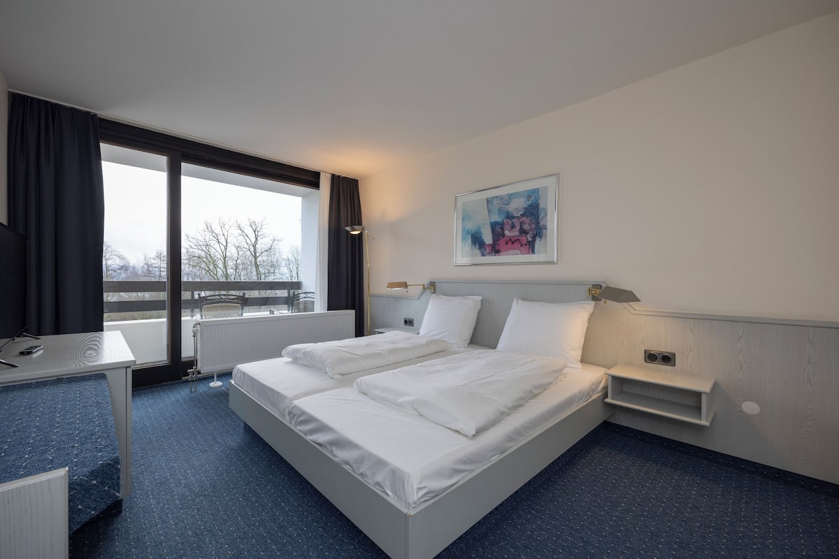 Une chambre d’hôtel avec un grand lit, un bureau, une chaise et une fenêtre donnant sur des arbres.