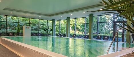 An indoor swimming pool with clear glass walls, a tiled floor, and a few lounge chairs along the edge.