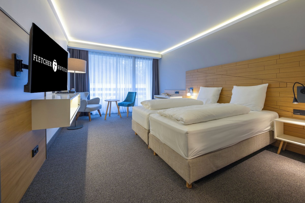 Une chambre d’hôtel équipée d’un téléviseur à écran plat, d’un bureau avec une chaise, de deux lits, d’une tête de lit en bois, d’une table de chevet avec une lampe et d’une fenêtre avec des rideaux.