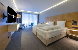 Habitación de hotel con televisión de pantalla plana, escritorio con silla, dos camas, cabecera de madera, mesita de noche con lámpara y ventana con cortinas.