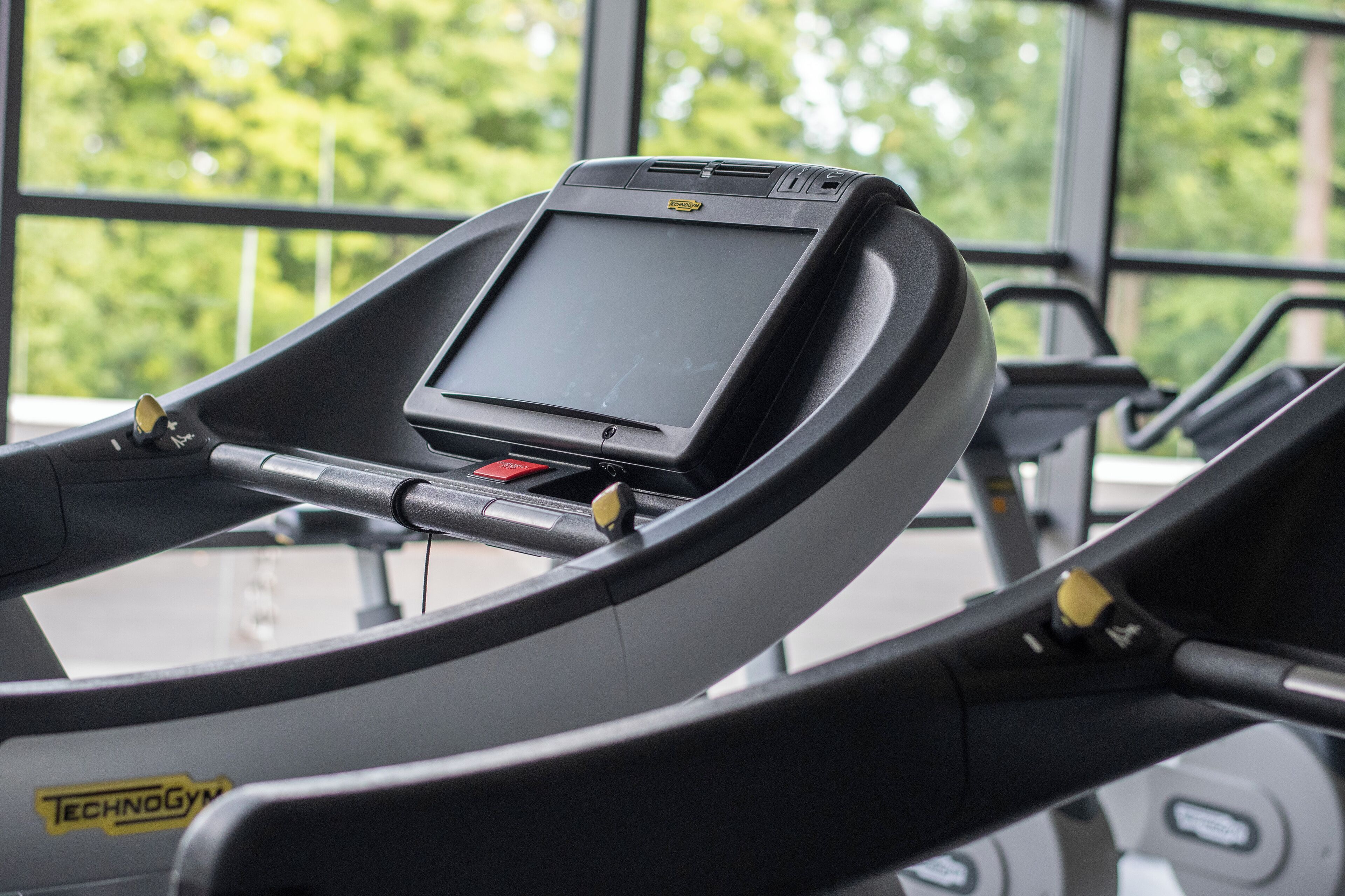 Una cinta de correr con pantalla digital y panel de control, ubicada frente a una ventana que da a vistas de vegetación.