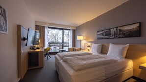 A hotel room with a large bed, a desk with a chair, a TV, and a window with a view of trees.