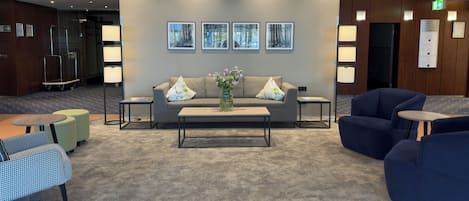 A lobby with a sofa, two side tables, a coffee table, and a vase with flowers. There are framed pictures on the wall and a glass elevator.