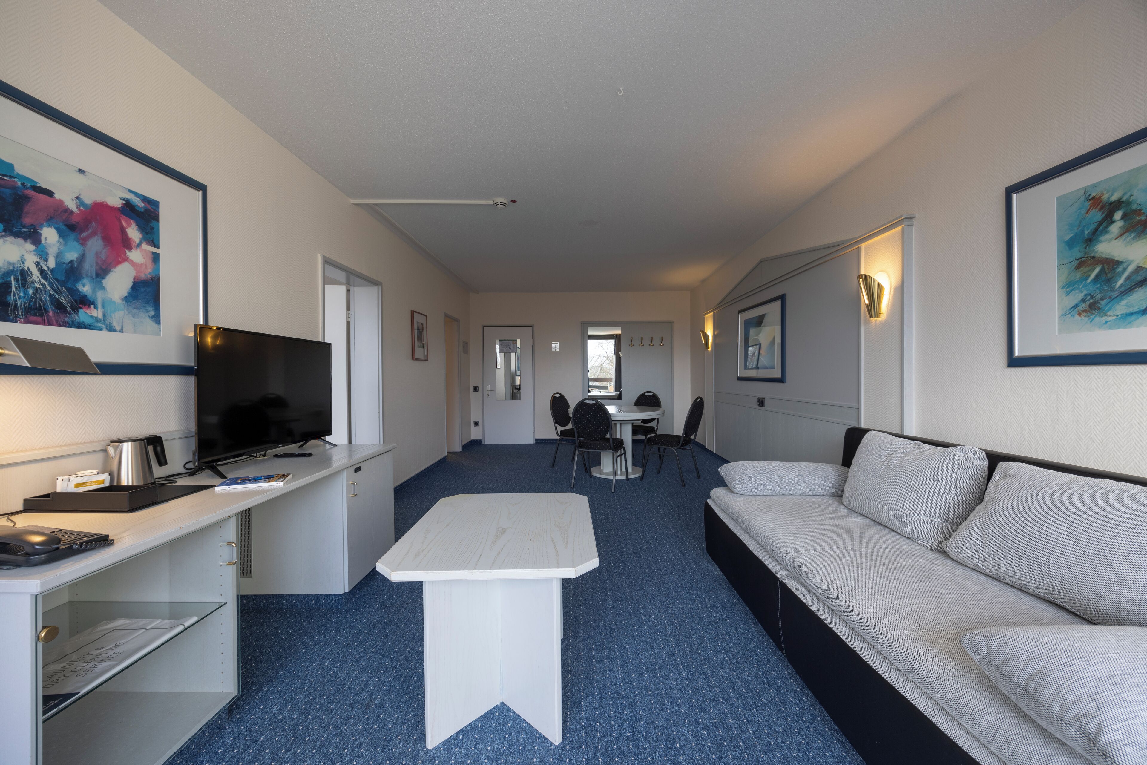 Habitación de hotel moderna con sofá, una mesita, escritorio con computadora, televisión y obras de arte en las paredes.